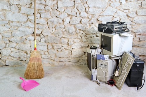 The image shows a collection of discarded electronic waste and household items arranged in front of a textured stone wall, likely in a garage or storage area. Prominent objects include a small CRT television with a curved screen, a desktop computer tower, an old beige computer monitor, a wired keyboard, a mouse, and a compact stereo system with visible cables. Toward the left side of the image, a pink plastic dustpan and a wooden-handled broom with yellow bristles are leaning against the stone wall. The floor surface appears concrete or similar material, and the scene is illuminated by natural or diffuse lighting, giving the setting a neutral appearance. This assortment of rubbish reflects typical household electronic waste stored temporarily before removal or disposal, highlighting the types of items that Garden Clearance Canary Wharf may assist in clearing from residential or commercial properties in the local area, including Canary Wharf or nearby districts within the postcode inferred from the webpage context.