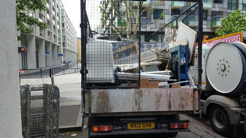 A flatbed truck parked on a city street, loaded with various discarded items including white panels, metal frames, and miscellaneous rubbish, all stacked haphazardly on the rear platform. The truck has a rusted metal base and is positioned near a modern residential building with light-colored exterior walls and balconies visible in the background. In the street setting, a traffic light and pedestrian crossing are seen, with minimal other traffic or pedestrians nearby. The environment suggests an urban area in Canary Wharf, with the company Garden Clearance Canary Wharf providing rubbish removal services in this location. The scene captures typical waste clearance activities, with the clutter of materials awaiting proper disposal or recycling, highlighting the company's role in professional rubbish collection within the area postcode and vicinity.