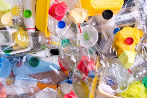 A close-up view of a pile of assorted plastic waste, including crushed and uncrushed bottles, bottle caps, and other small container fragments. The plastics vary in colour from transparent, yellow, red, green, to blue, with some items featuring visible labels or branding. The objects are scattered chaotically, with some bottles partially squeezed or flattened, showing different textures such as glossy, matte, and crinkled surfaces. The background shows more plastic debris, creating an overall impression of collected waste suitable for rubbish removal services, particularly in an urban or garden clearance context nearby Canary Wharf. The arrangement emphasizes the variety of plastic materials and colours typical of waste collected from household or commercial sources, highlighting the necessity for professional rubbish clearance to manage such refuse efficiently, consistent with services provided by Garden Clearance Canary Wharf.