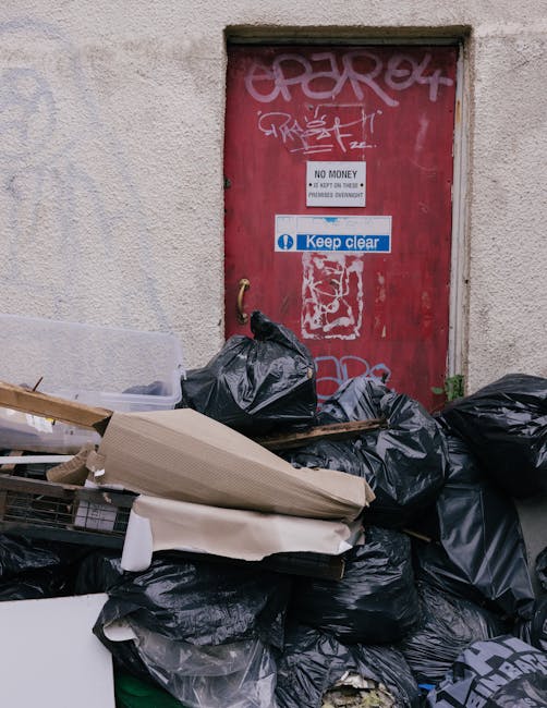A man sitting on the ground next to large plastic bags filled with various types of rubbish, positioned in front of a weathered, corrugated metal shutter door set in an urban environment. The bags are