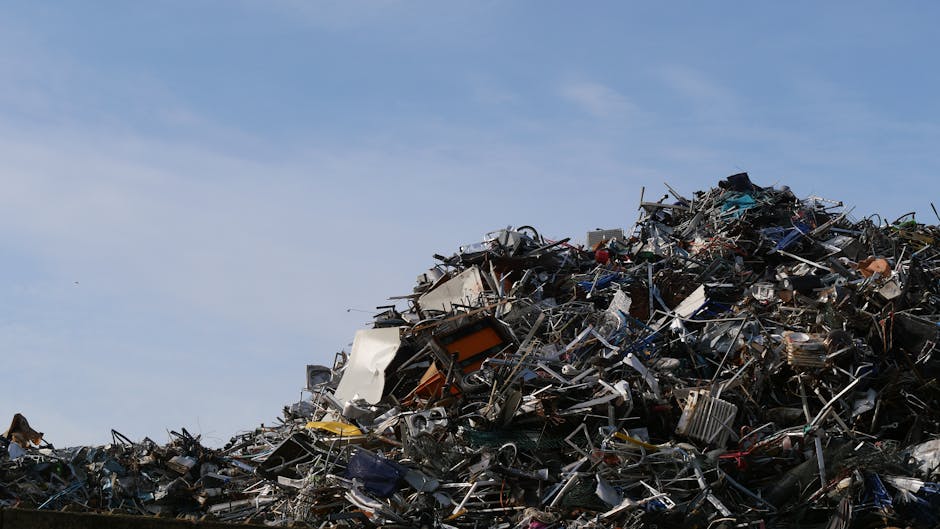 A large pile of mixed metal and electronic waste located outdoors under a clear blue sky. The heap contains various items such as discarded appliances, metal frames, cables, and scrap metal pieces, wi