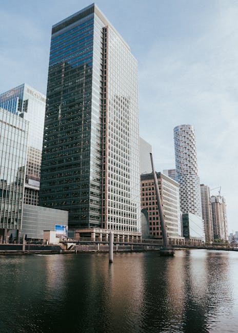 A modern urban scene featuring a low-angle view of high-rise office buildings with glass facades reflecting the overcast sky, located along a canal or river. In the foreground, there is a distinctive 